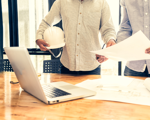 Man in button down shirt holding construction hat with laptop on table in front of him